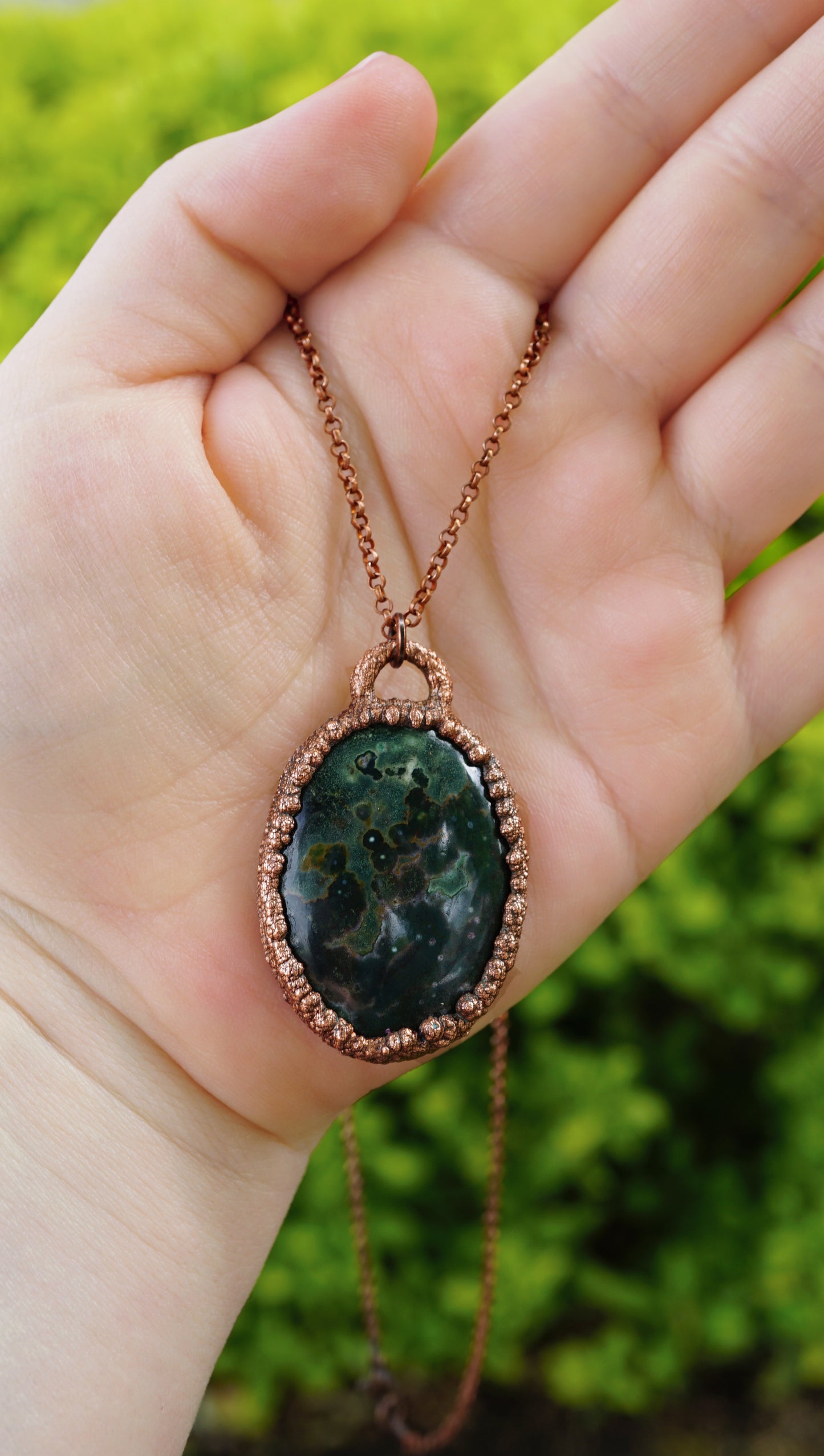 Hand holding a necklace with a green pendant against a blurred green background