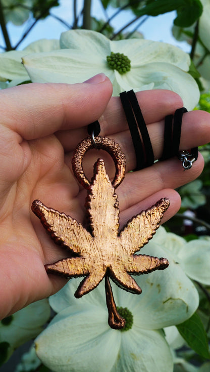 hand holding backside of copper ganja leaf pendant with black suede, against white dogwood flowers