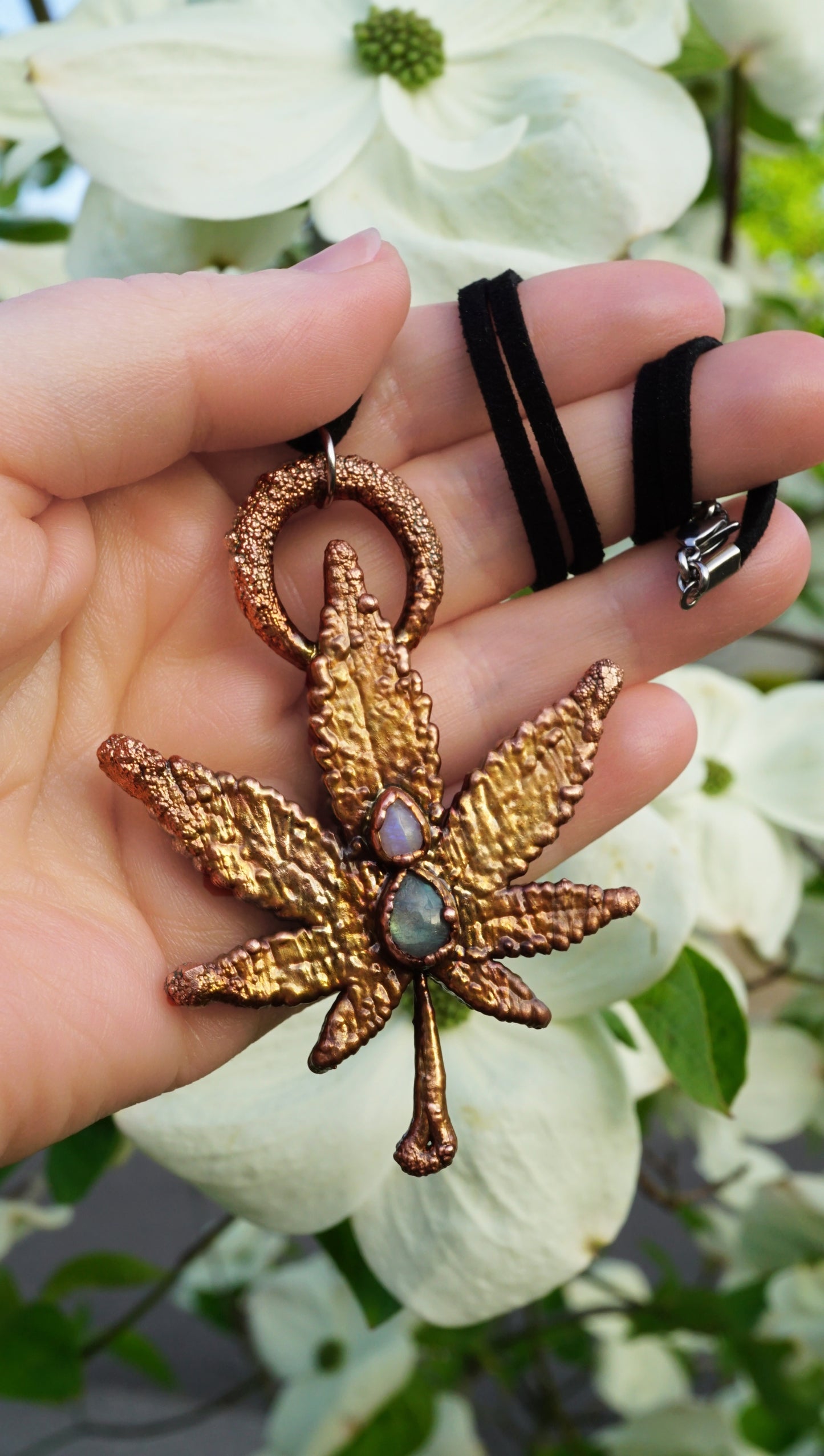 Hand holding a decorative leaf-shaped ornament with a gemstone, against a blurred floral background.