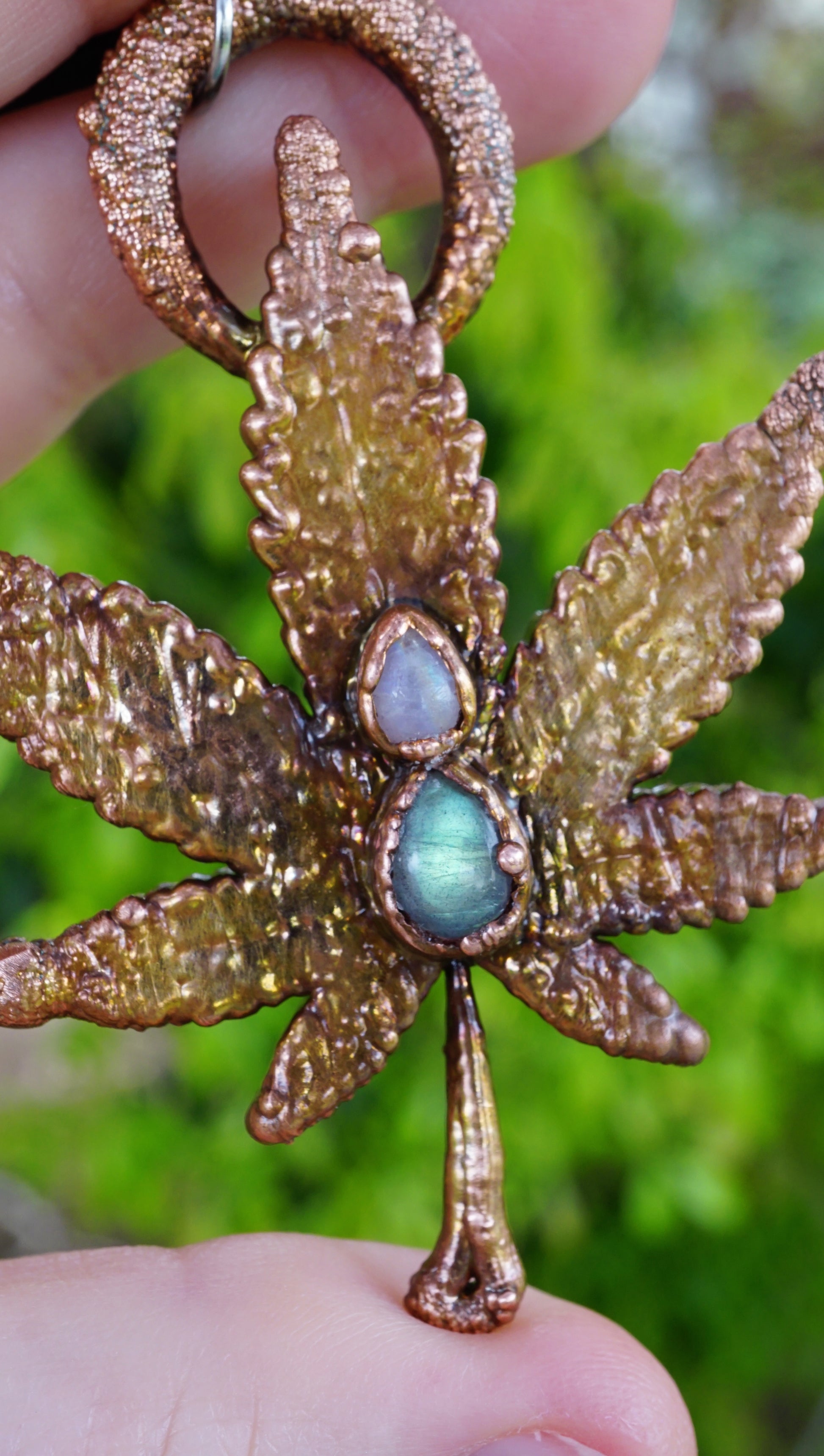 up close picture of pretty weed leaf necklace, featuring green and white gemstones. held in hand against greenery