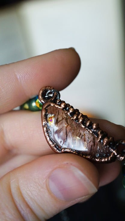 Copper and quartz necklace with a pendant held between fingers against a blurred background