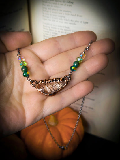 hand holding handmade necklace, green sparkly beads surrounding quartz crystal in copper. steel chain dangles around hand, with old book and mini pumpkin in background.