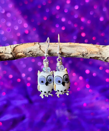 white Owl bead earrings with dark periwinkle purple eyes, hanging on a branch with a purple sparkly background