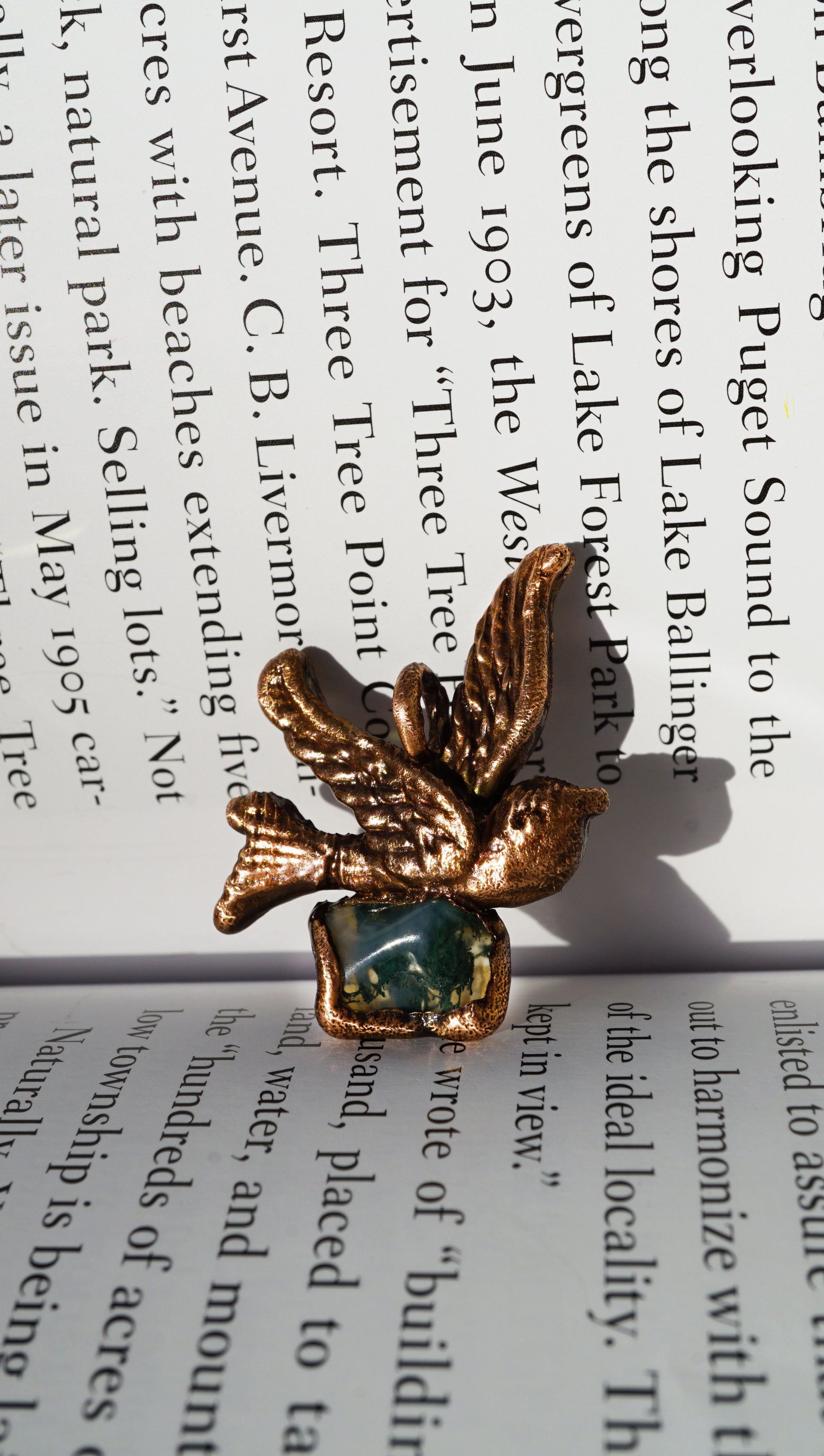 copper sparrow pendant holding a green mossy stone, against book page
