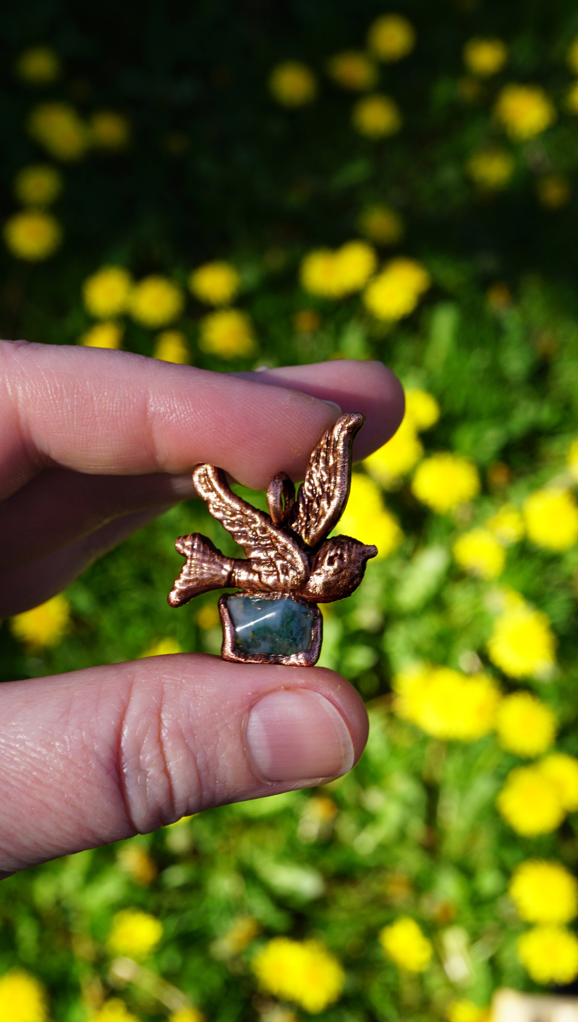 Copper-colored bird-shaped pendant setting with green moss agate that looks like a nest below the sparrow, held between fingers with a blurred background with yellow flowers and green grass