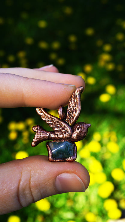 copper bird-shaped pendant with a green gemstone held between two fingers against a blurred green grass and yellow flowers