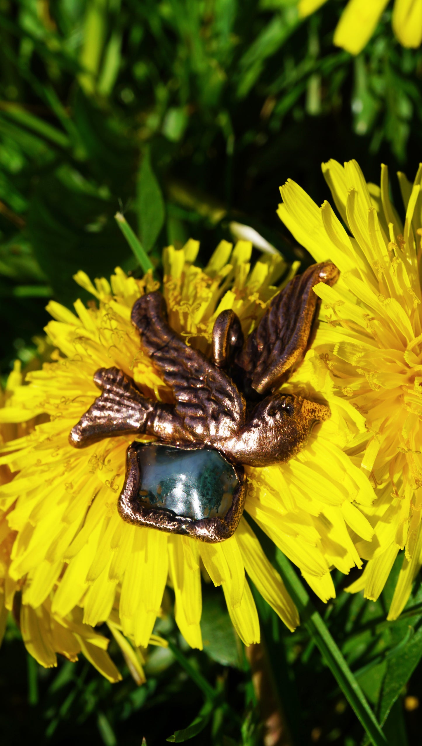antique coppery sparrow pendant with green mossy stone, laying on top of yellow dandelion flower and green grass, facing to the sky