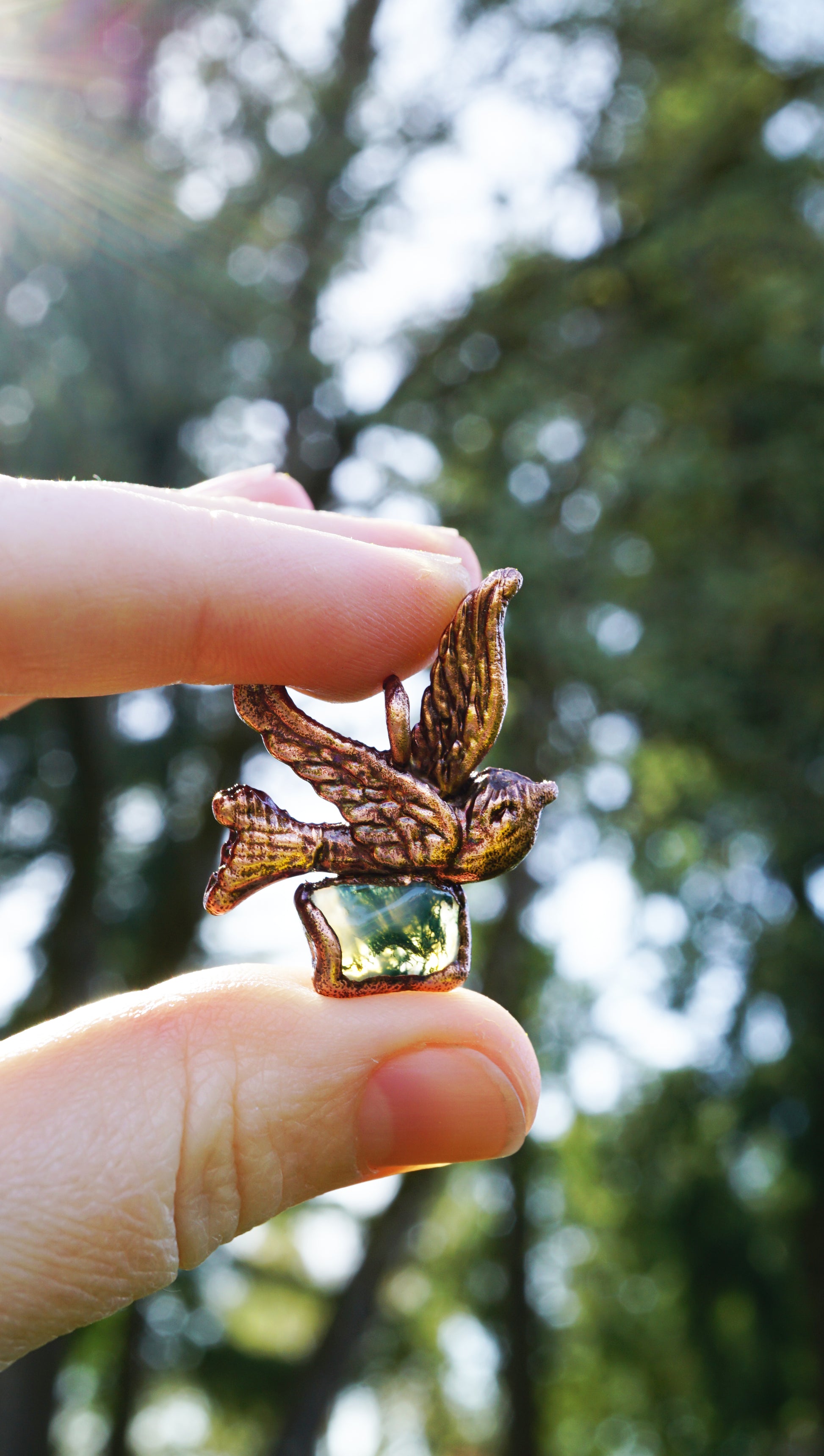 holding pendant between fingers in the sun against green trees and blue sky, pendant is an antiqued copper with green mossy stone