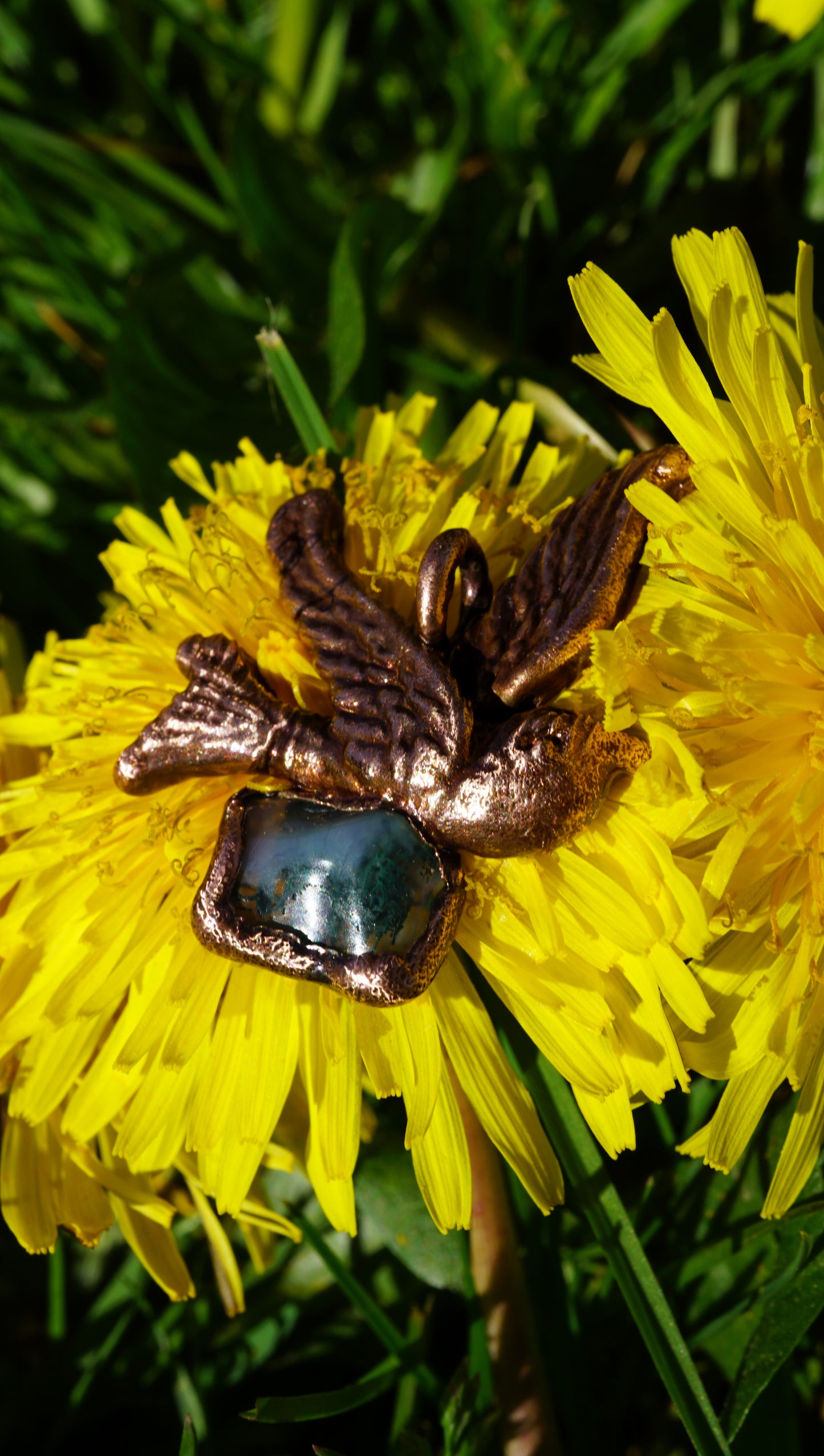 antique coppery sparrow pendant with green mossy stone, laying on top of yellow dandelion flower and green grass