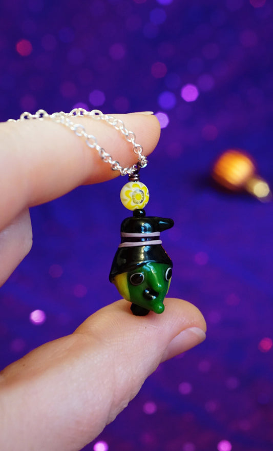 A hand holding a silver chain necklace with a small green witch head pendant. she has a black hat with a yellow sun bead above it, and matching bright yellow hair. her head is shaped like a crescent moon. set against a purple background.