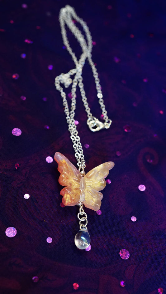 A silver chain necklace featuring a carved butterfly pendant made of translucent peach and orange stone, with a small clear bead charm, displayed on a dark purple glittery background.