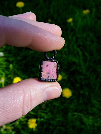 pink stone pendant with black copper setting, rectangular shape. held in fingers against green grass and yellow flower background