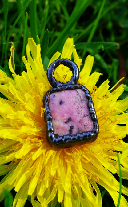 pink stone pendant with black copper setting, rectangular shape.  sitting on yellow flower against grass