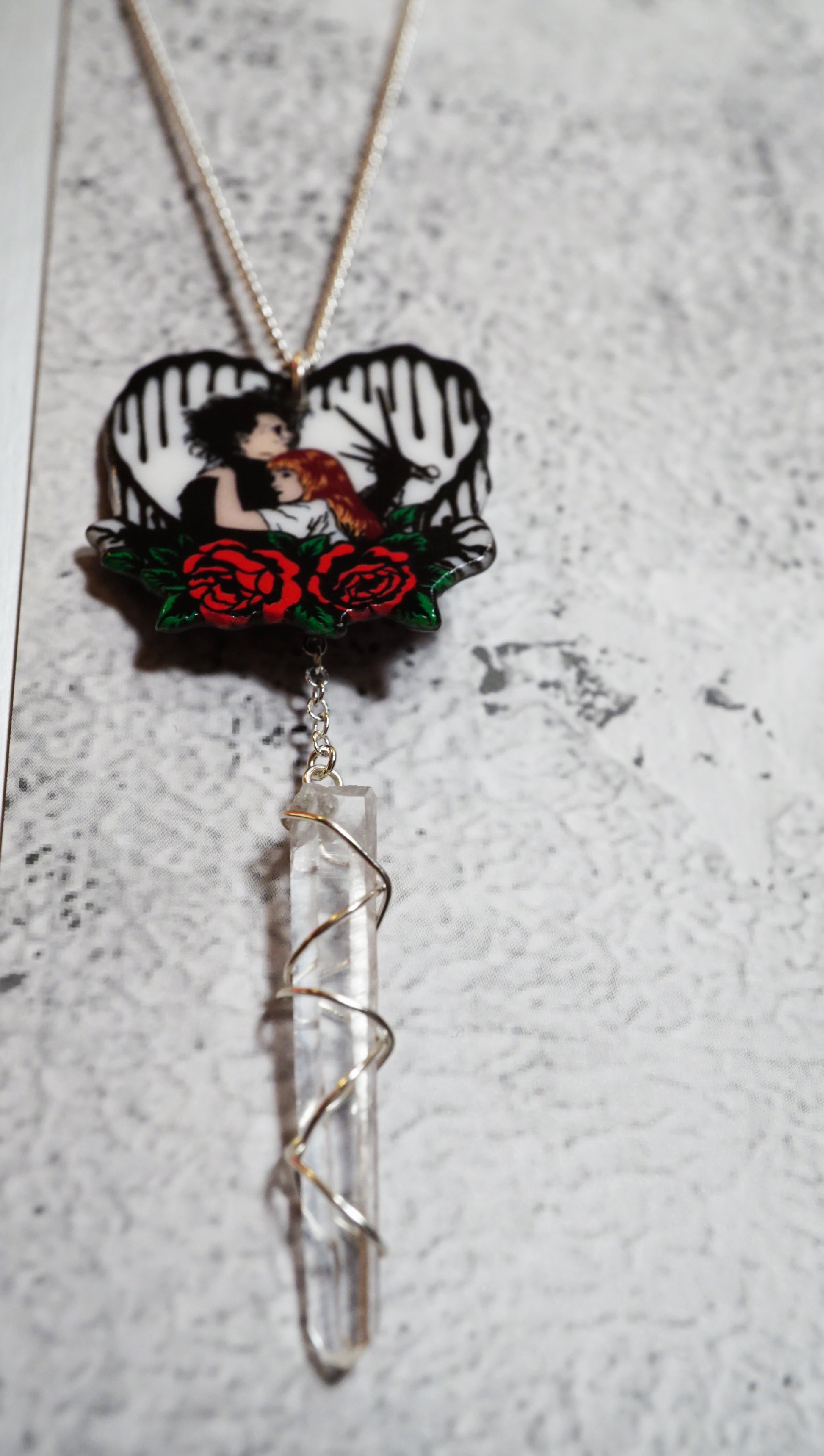 heart shaped charm necklace with wire wrapped clear quartz dangling below. red roses and edward scissorhands holding him are on the charm. white snowy background. 