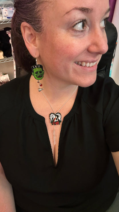 woman wearing creepmas jewelry. frankenstein's monster earrings with steel bolts, and edward scissorhands heart necklace with clear quartz. wearing black shirt. 