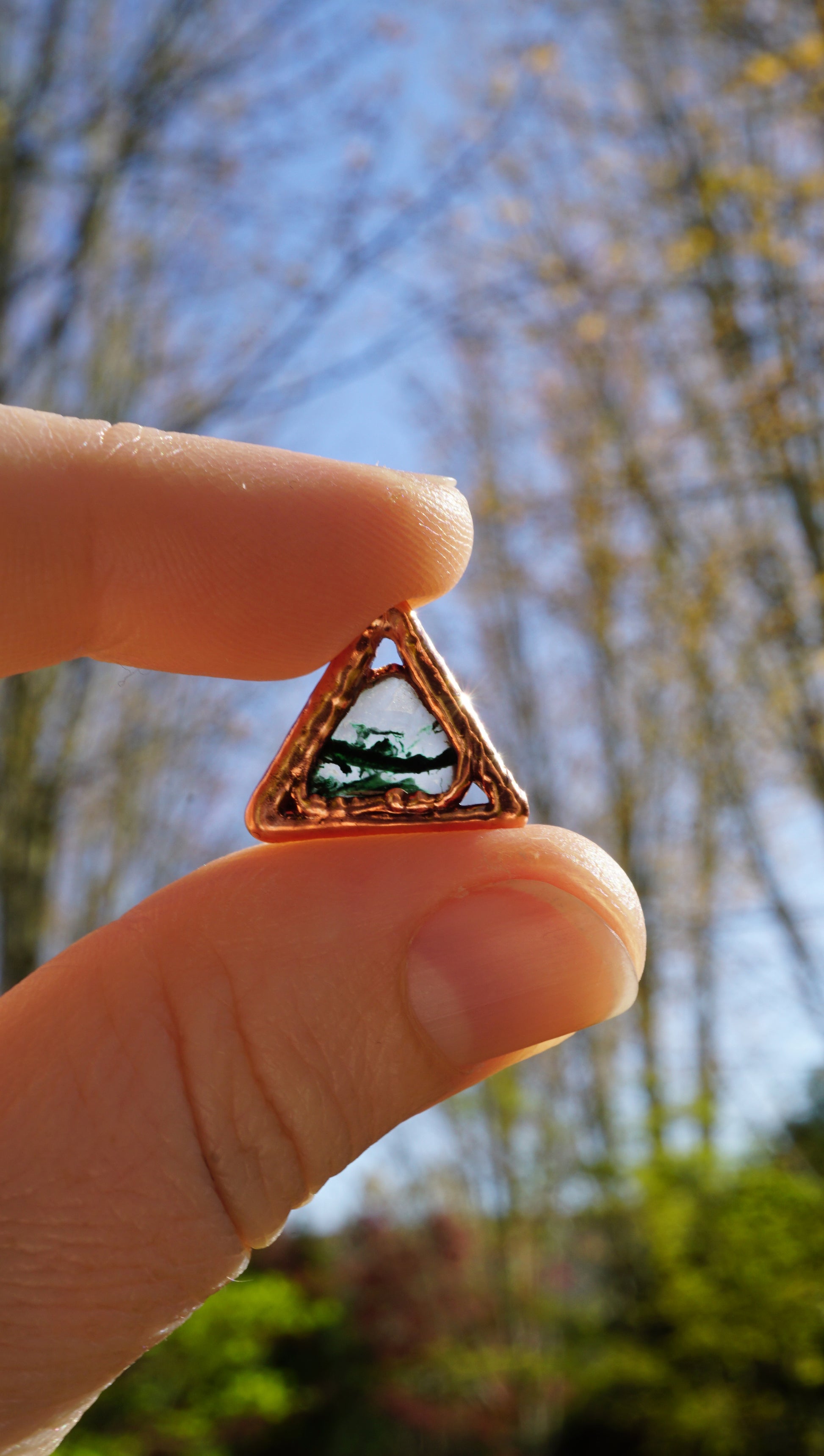 Hand holding a small triangular copper charm with dark green and clear stone in center, with a blurred natural background outside in the sun