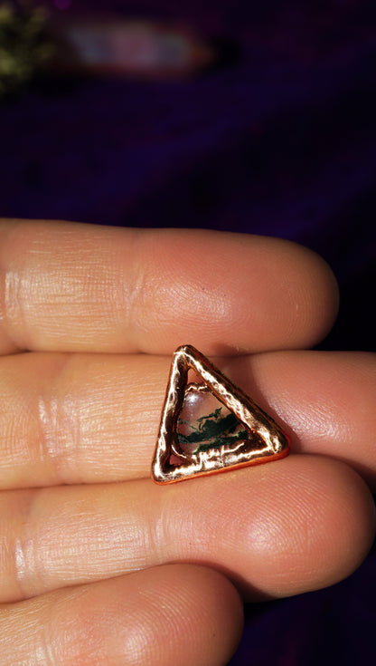 Rose gold triangle-shaped necklace charm on a finger against a blurred purple background