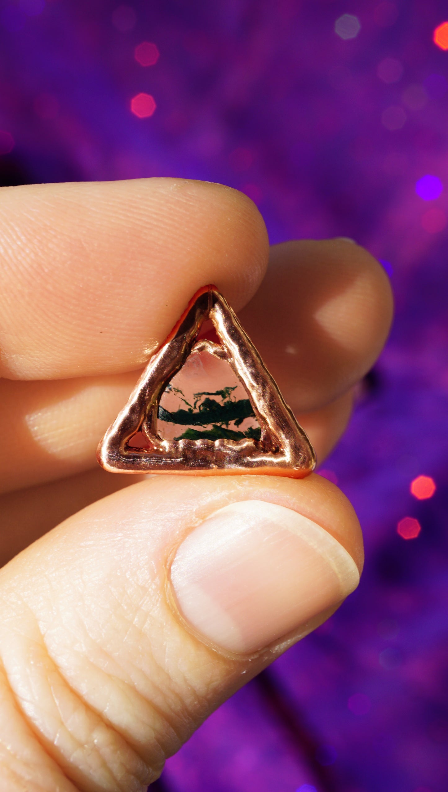 handmade electroformed copper triangle charm with dark green stone, purple background, held in fingers