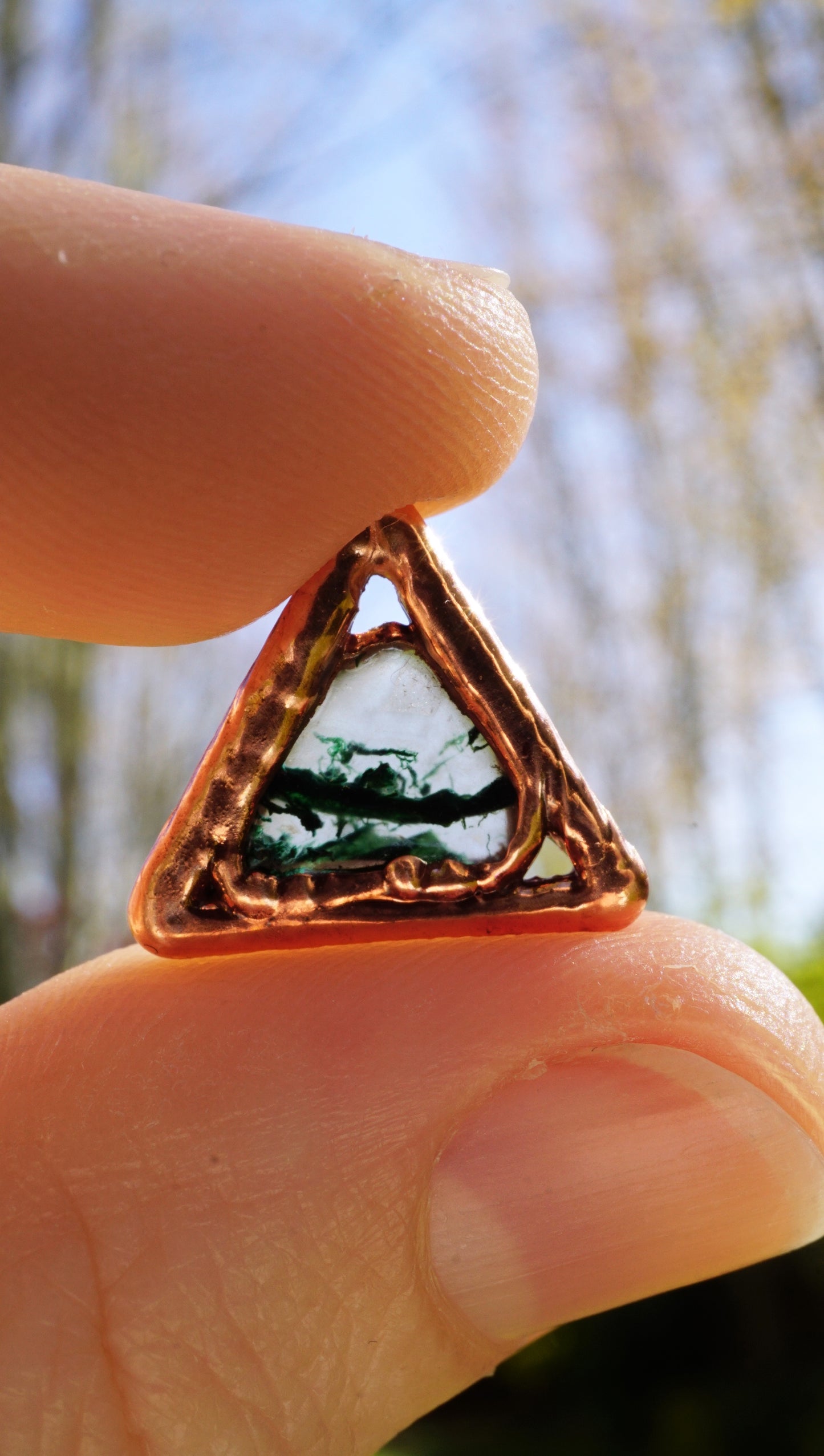 triangular shaped copper pendant with green and clear moss agate stone in center, held between fingers in sun