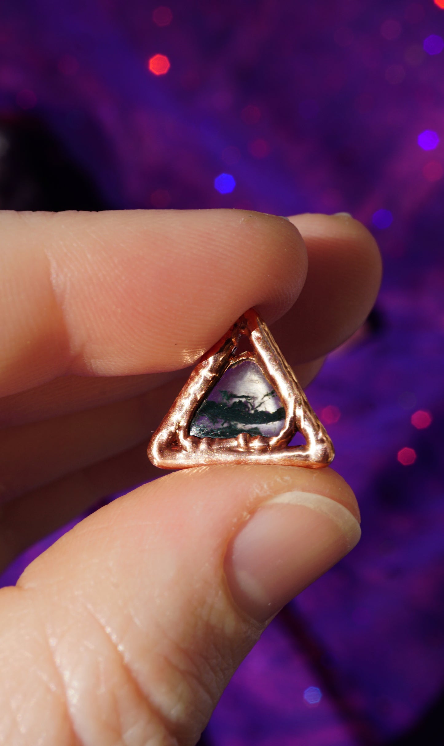 triangle pendant between fingers, it glows in the sun with purple background. dark green stone in center, looks like moss