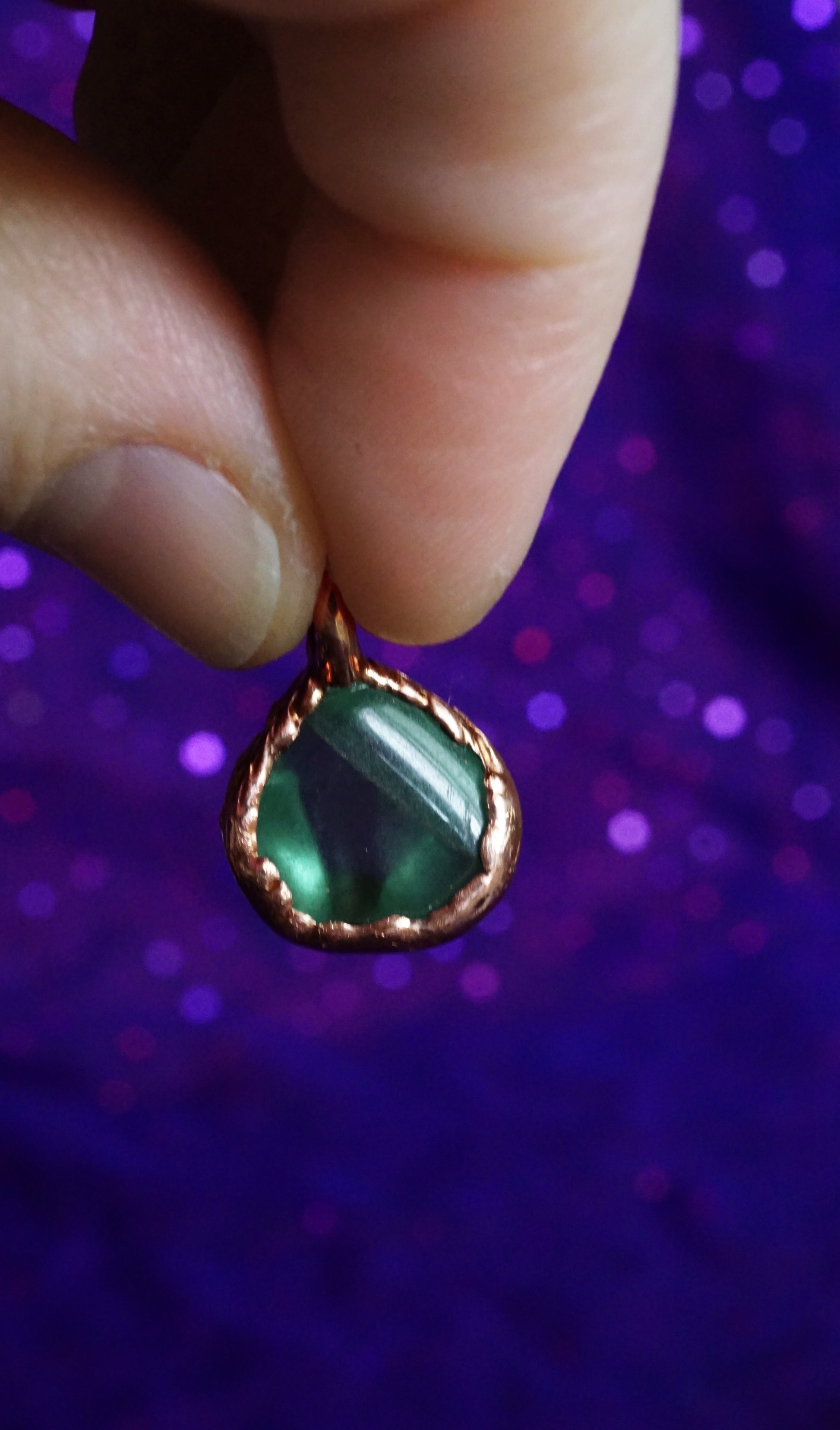 backside of copper pendant with green stone. natural crack in stone showing. in between fingers, purple fabric background 