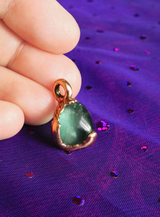 Green stone pendant with light copper setting held by a hand on a purple fabric background