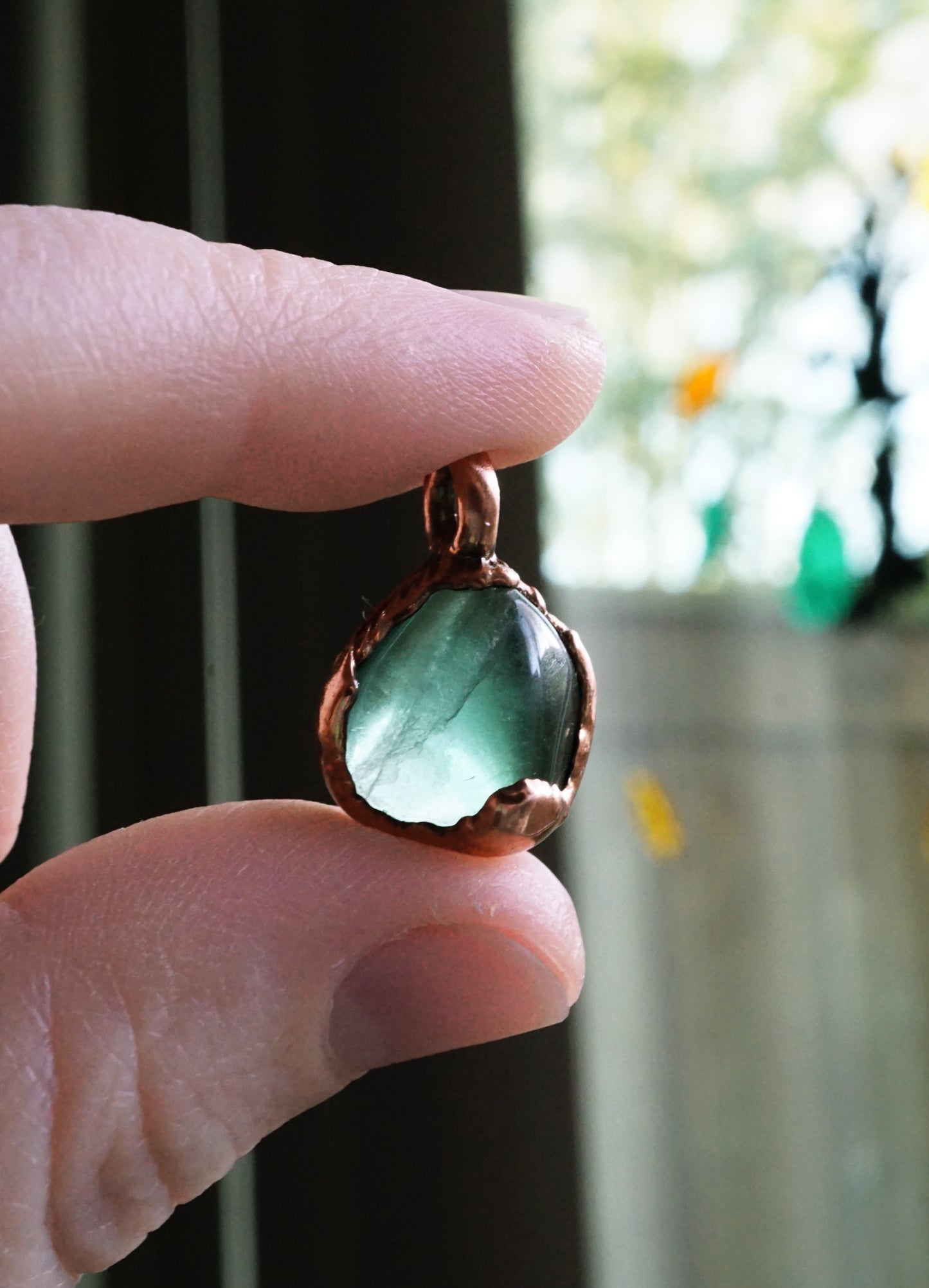 Hand holding a pendant with a thick green teardrop-shaped gemstone against a blurred outdoor background.