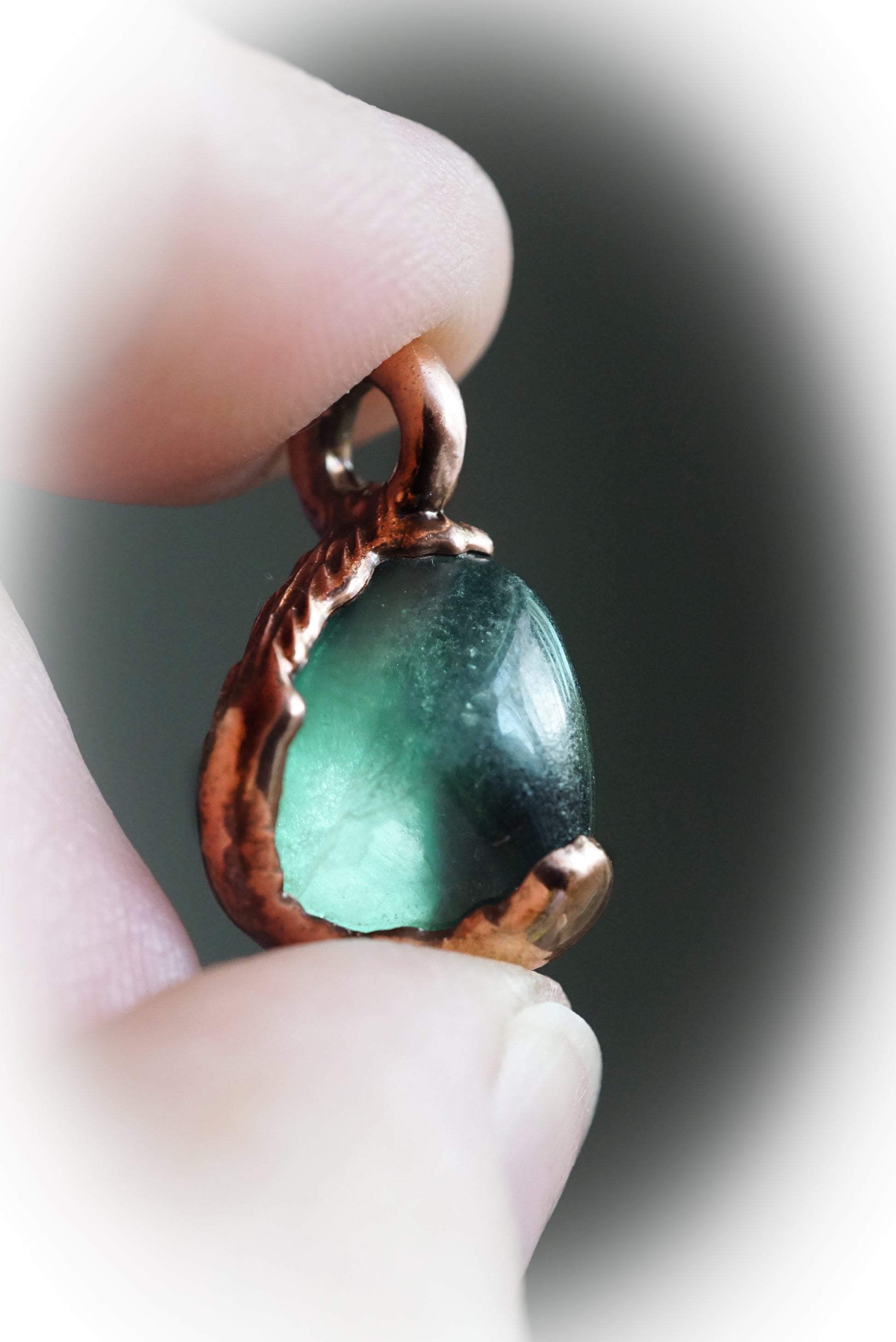 Green crystal pendant with copper setting held between fingers against a blurred background