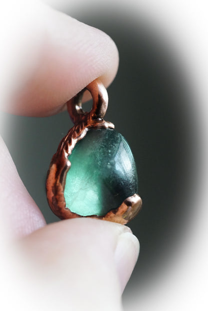 Green crystal pendant with copper setting held between fingers against a blurred background
