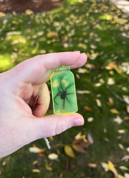 Hand holding a small transparent greenish yellow jar necklace with a long legged black spider image inside, against a blurred natural background
