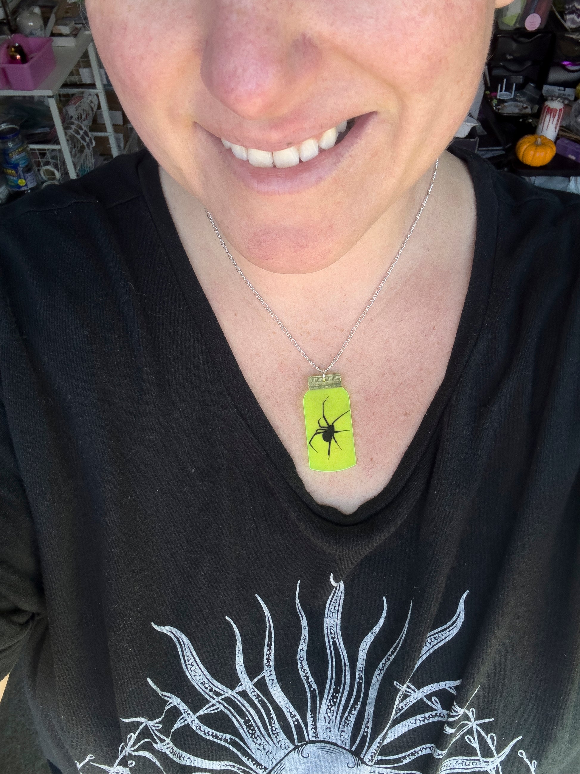 Person wearing a black shirt with a graphic design, smiling. wearing a mountain dew-yellow jar necklace with black spider image with long legs