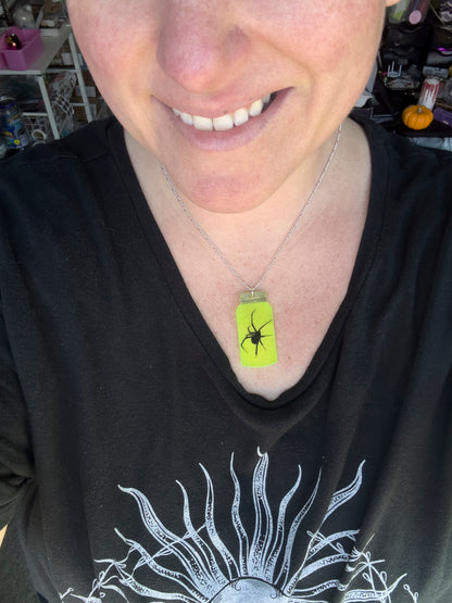 Person wearing a black shirt with a graphic design, smiling. wearing a mountain dew-yellow jar necklace with black spider image with long legs