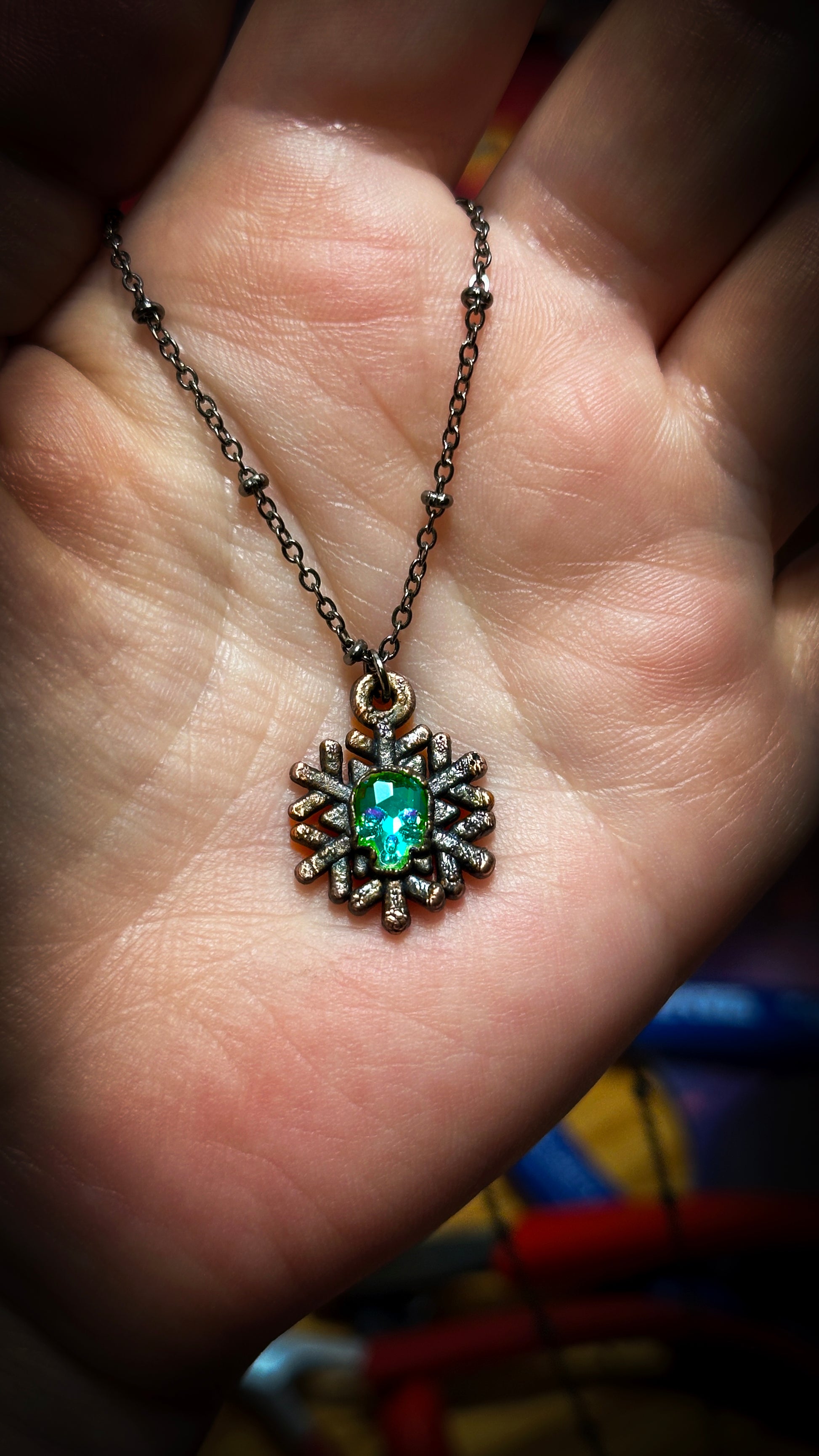 copper snowflake necklace with shiny green skull rhinestone in the center. strung on hand with gunmetal chain. jewelry tools behind that. 