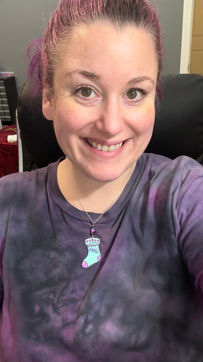 Person wearing a purple shirt with a necklace featuring a cute but goth stocking, in mint with pink accents, a tiny lavender bat and skulls. purple and black beads at top. silver chain.
