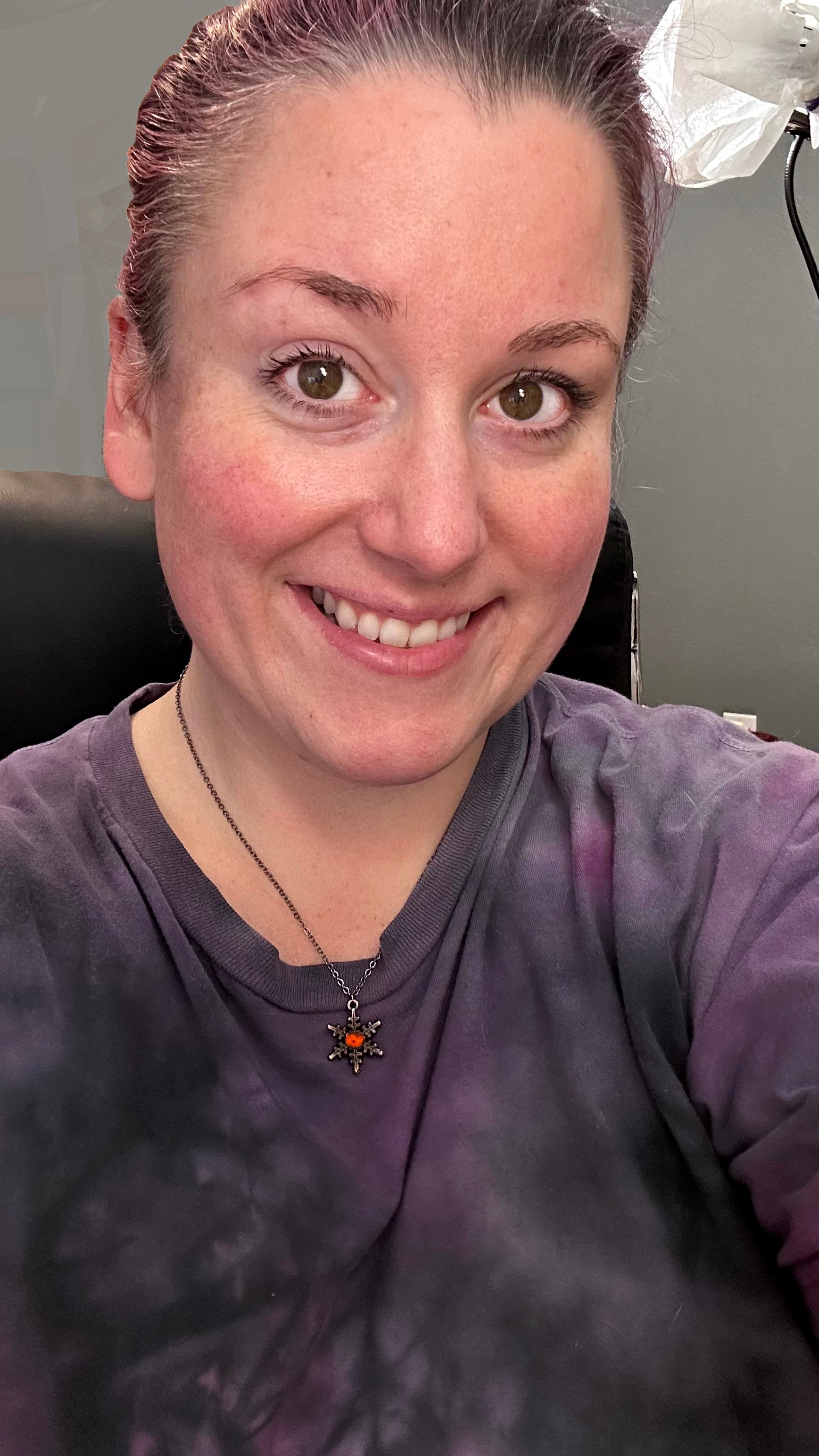Person wearing a purple shirt with a jack o lantern snowflake necklace. sitting in an office chair.